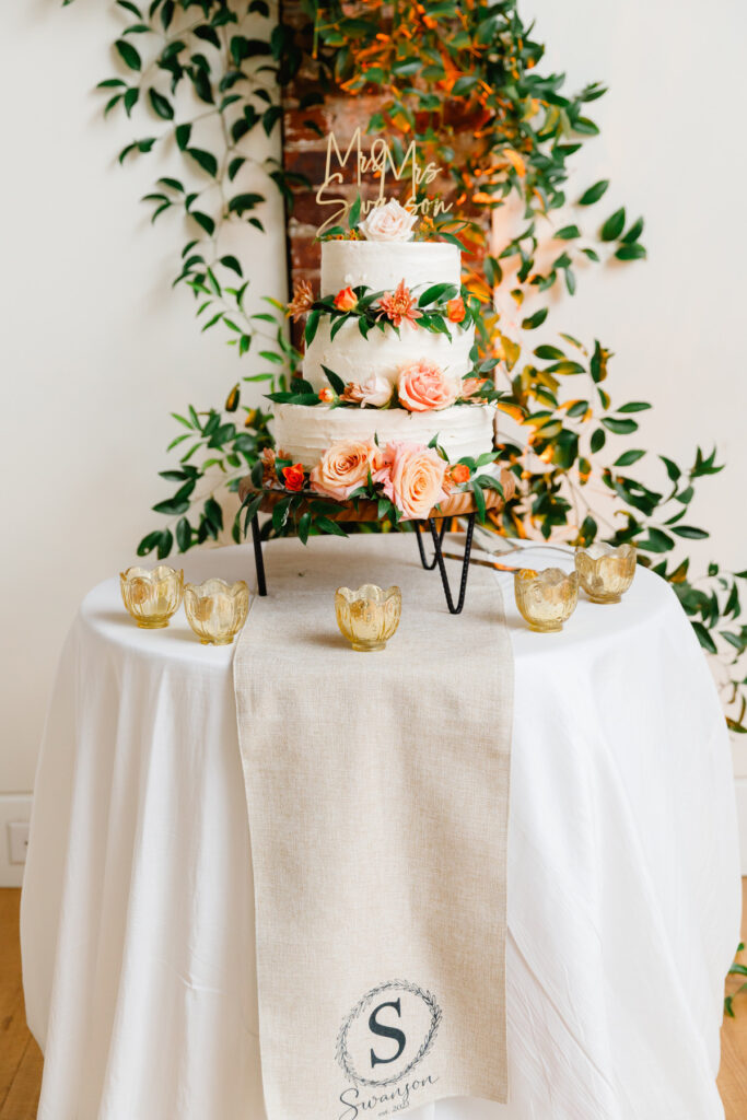 Three tiered all white wedding cake with orange and pink roses and a gold Mr Mrs Swanson cake topper