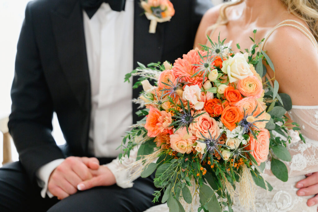 Photo of bride and groom holding hands with focus on bridal bouquet with orange and pink roses and blue flowers