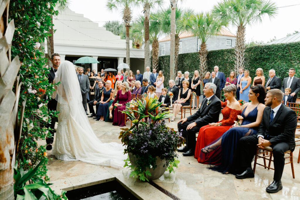 View of wedding guests during ceremony seated in chairs and standing in the courtyard at a Cannon Green Wedding