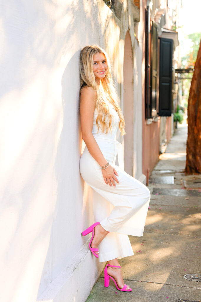 A young woman with blond hair wearing a strapless white jumpsuit and hot pink high heels standing in front of a white historic house in downtown Charleston, SC