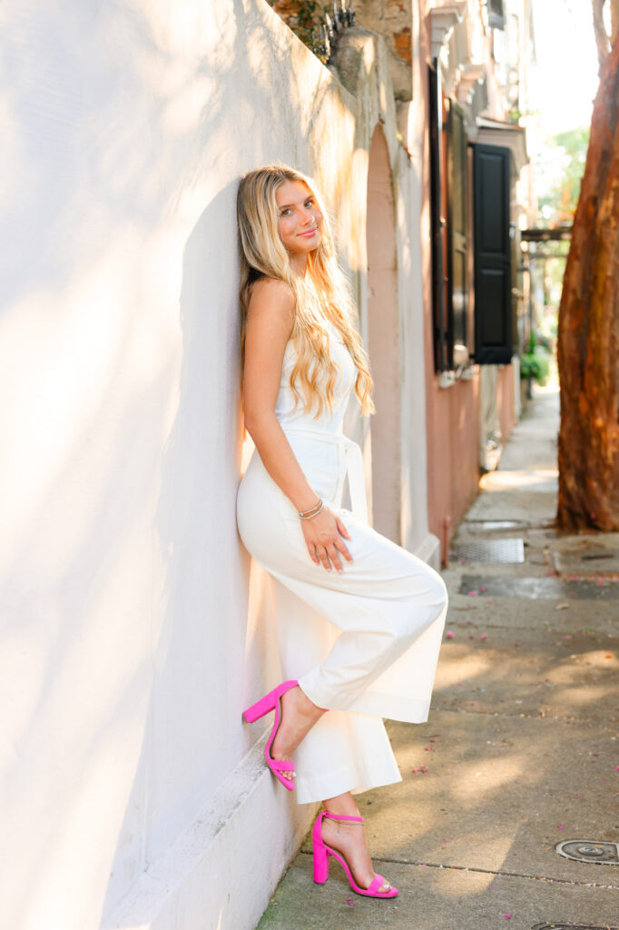 A young woman with blond hair wearing a strapless white jumpsuit and hot pink high heels standing in front of a white historic house in downtown Charleston, SC