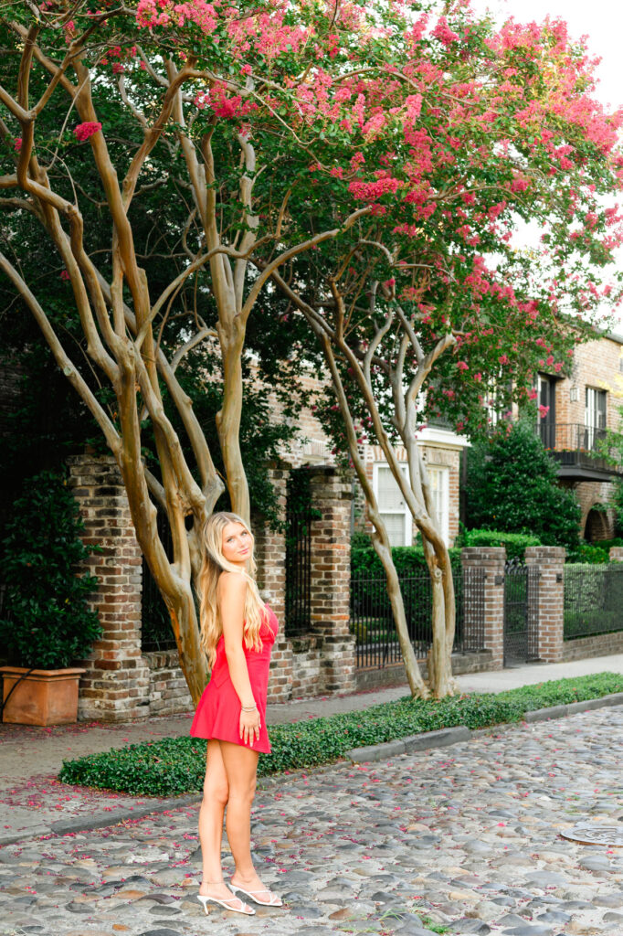 A young woman with blond hair wearing a strapless red dress with a bow in the back standing on a cobblestone street in historic downtown Charleston, SC on S Adgers Wharf