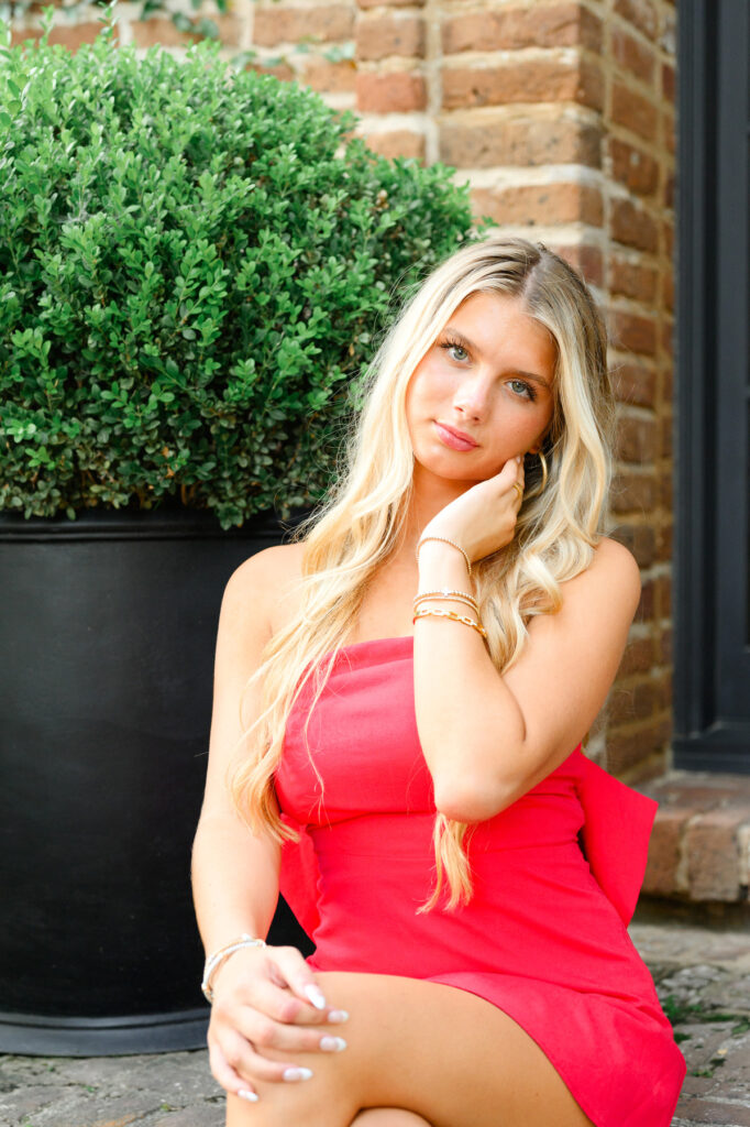 A young woman with blond hair wearing a strapless red dress with a bow in the back sitting on steps in historic downtown Charleston, SC, on S Adgers Wharf