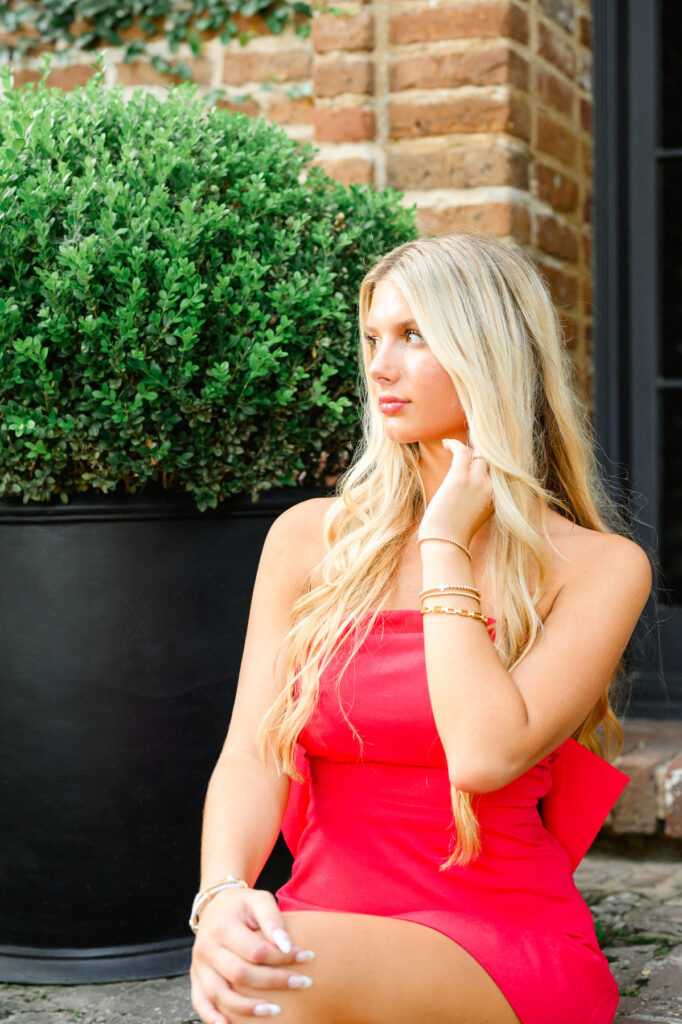 A young woman with blond hair wearing a strapless red dress with a bow in the back sitting on steps in historic downtown Charleston, SC, on S Adgers Wharf