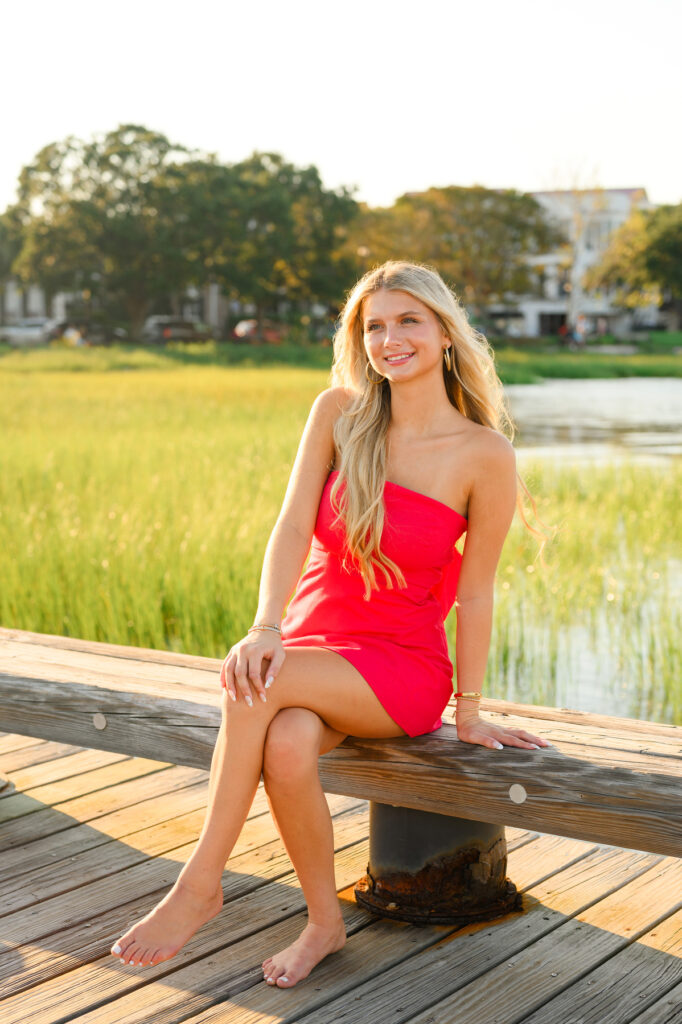 A young woman with blond hair wearing a strapless red dress with a bow in the back in historic downtown Charleston, SC, at Waterfront Park sitting on pier