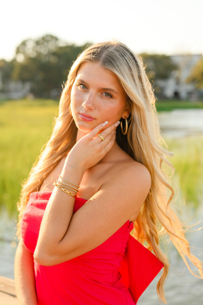 A young woman with blond hair wearing a strapless red dress with a bow in the back in historic downtown Charleston, SC, at Waterfront Park sitting on pier