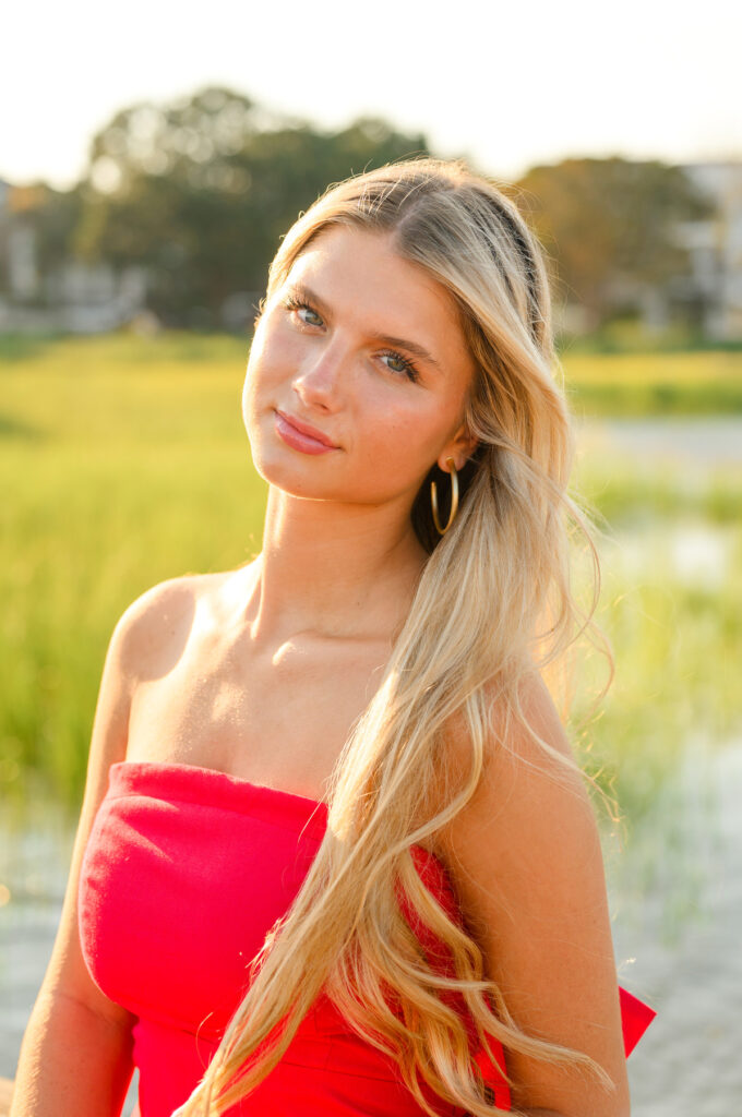 A young woman with blond hair wearing a strapless red dress with a bow in the back in historic downtown Charleston, SC, at Waterfront Park sitting on pier