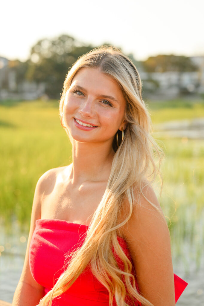 A young woman with blond hair wearing a strapless red dress with a bow in the back in historic downtown Charleston, SC, at Waterfront Park sitting on pier