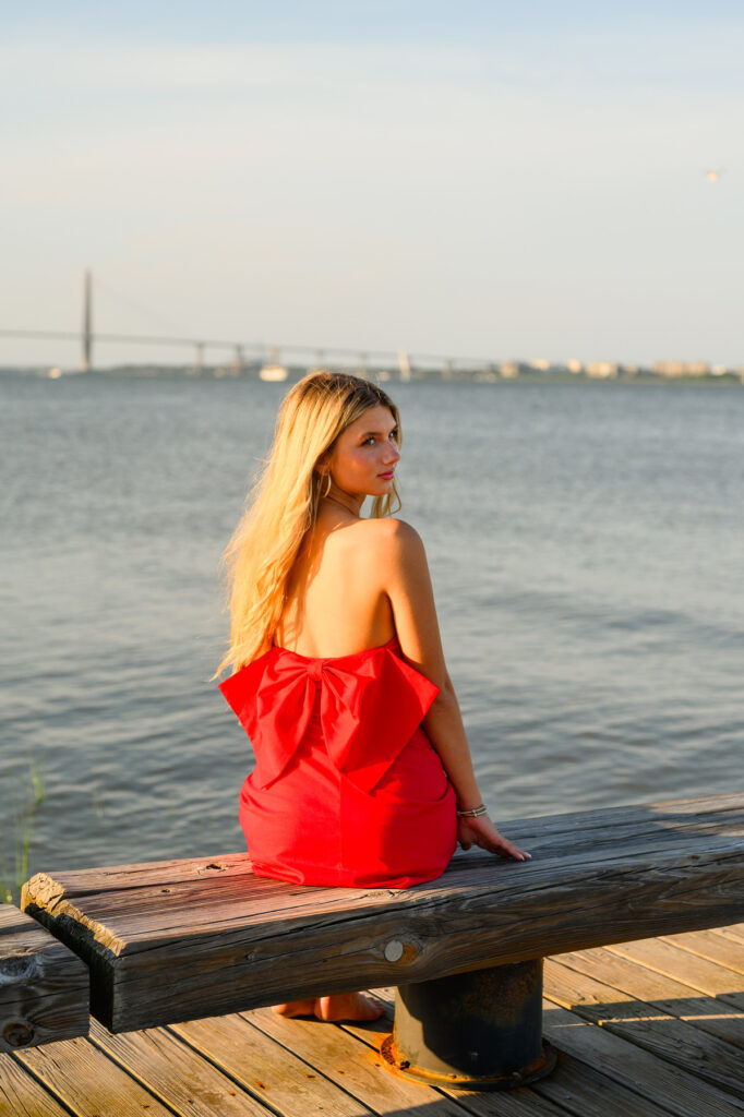 A young woman with blond hair wearing a strapless red dress with a bow in the back in historic downtown Charleston, SC, at Waterfront Park sitting on pier