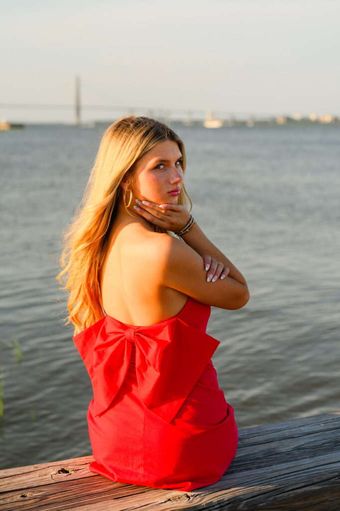 A young woman with blond hair wearing a strapless red dress with a bow in the back in historic downtown Charleston, SC, at Waterfront Park sitting on pier