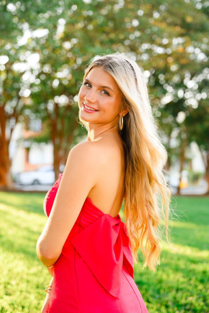 A young woman with blond hair wearing a strapless red dress with a bow in the back in historic downtown Charleston, SC, at Waterfront Park