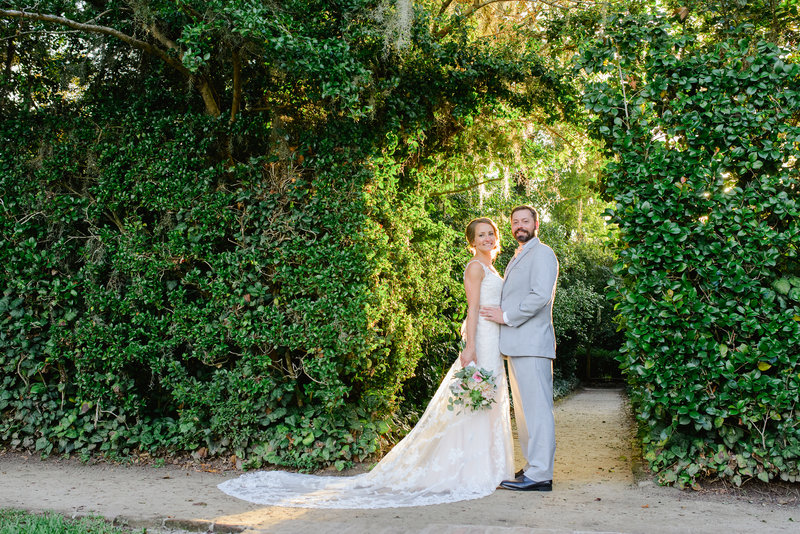 A bride in a white lace dress and a groom in a light gray suit pose for a portrait during their Middleton Place wedding.