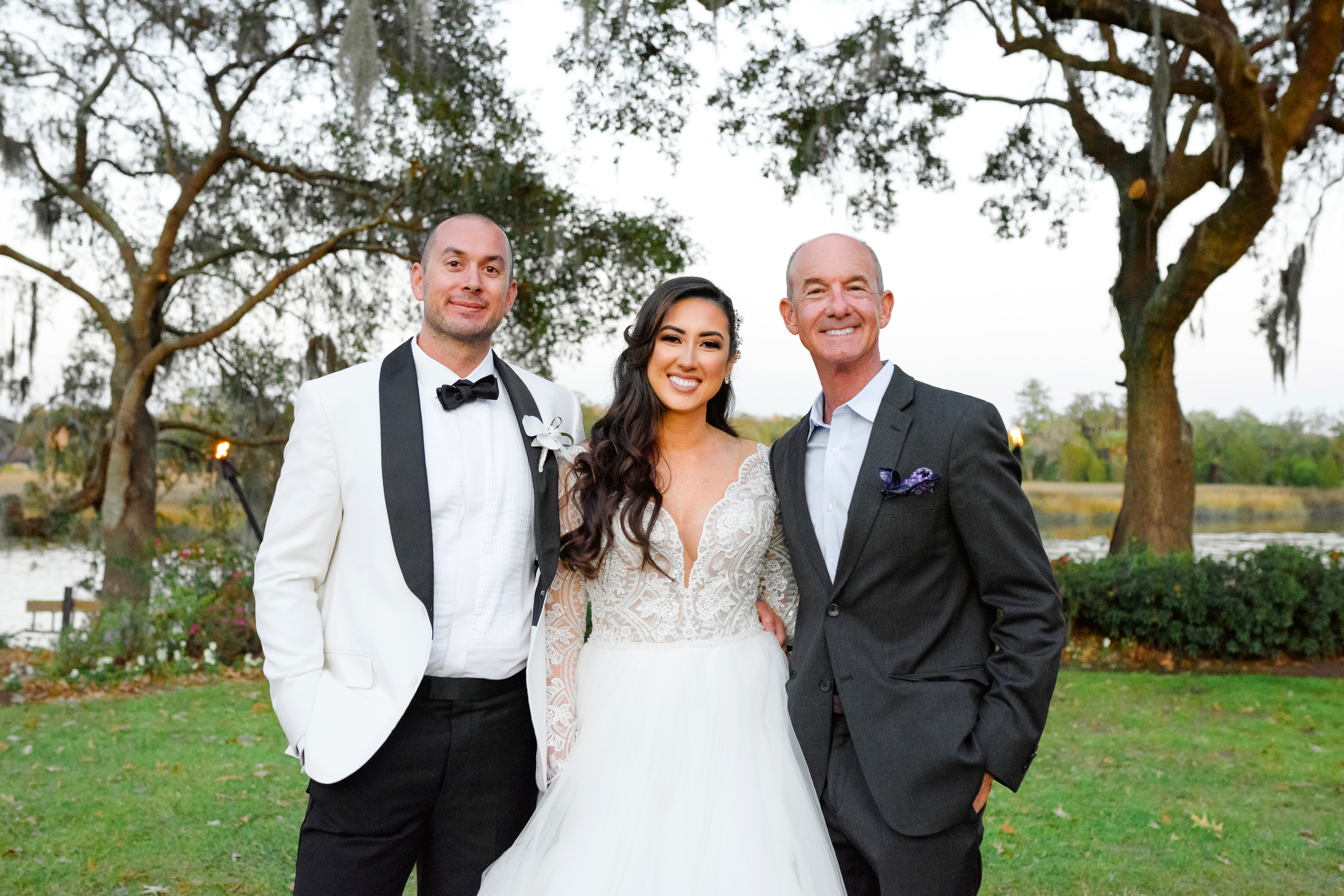 Charleston wedding planner Mike Winship poses for a portrait with a groom in a white tux with black pants and a bride in a white dress.