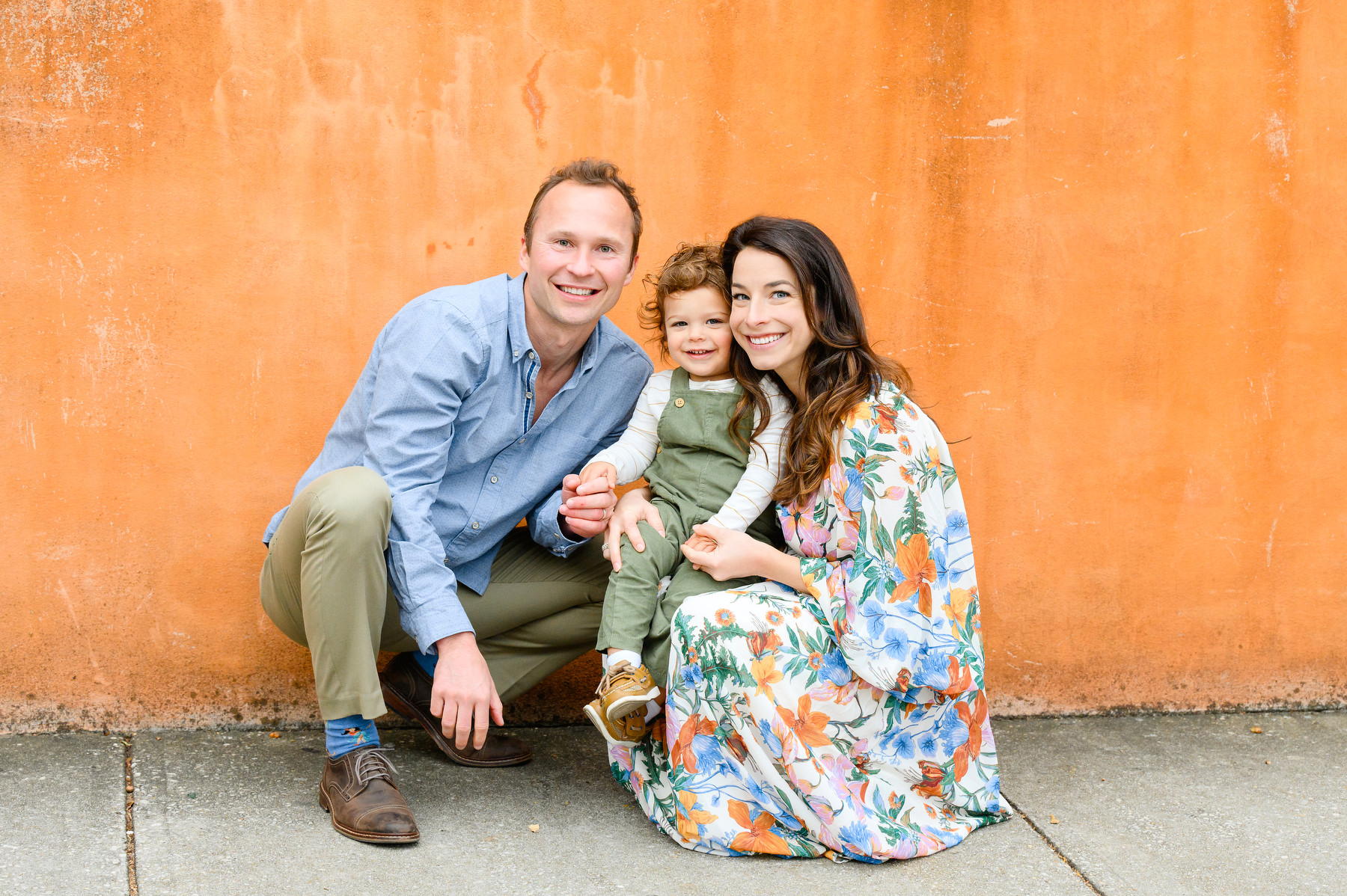 Downtown Charleston family photos with a father in a blue shirt, toddler in green overalls, and a mother in a floral print maxi dress