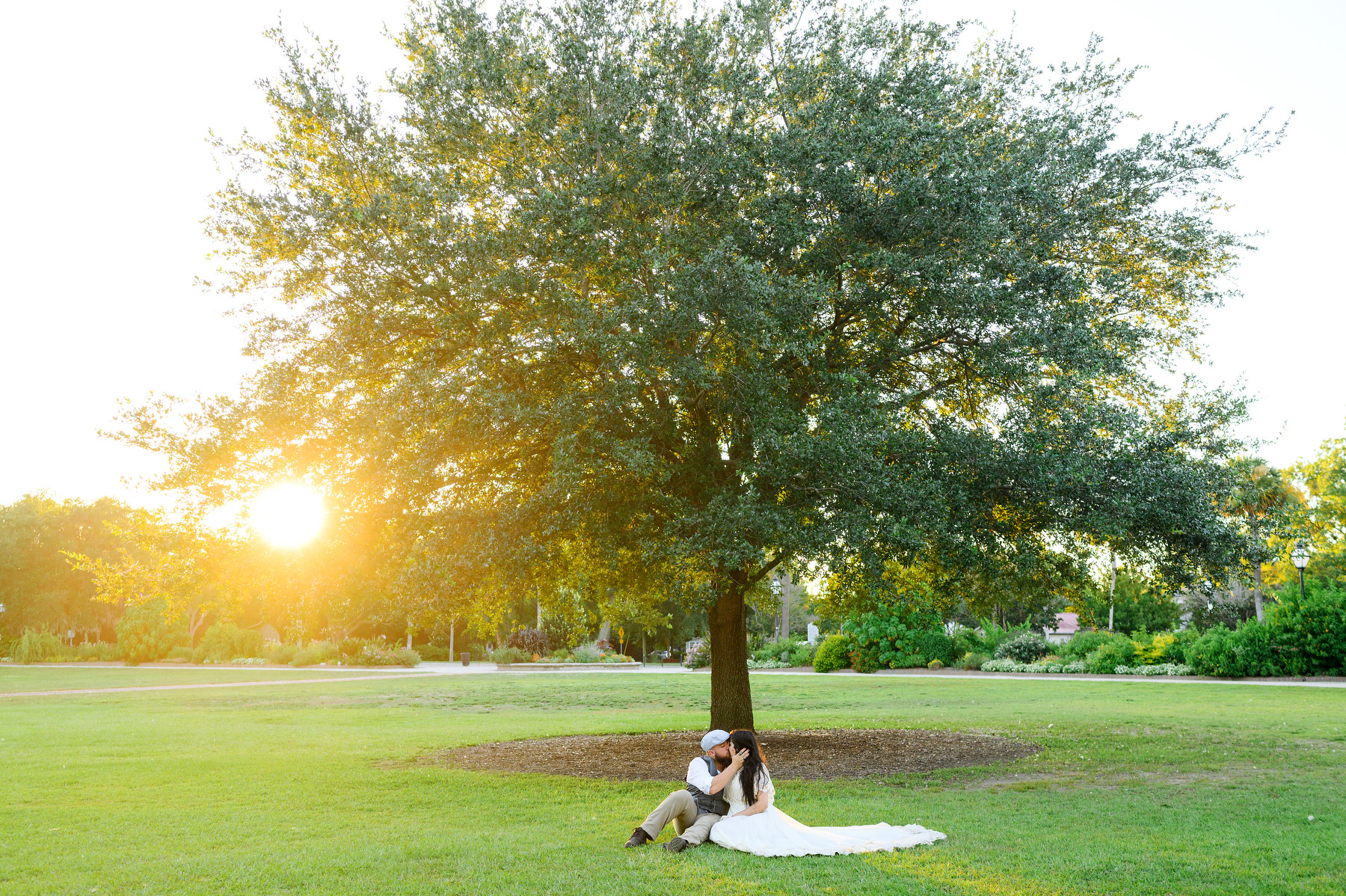 A man and a woman sit on the grass in front of a tree at Hampton Park during their engagement photos.